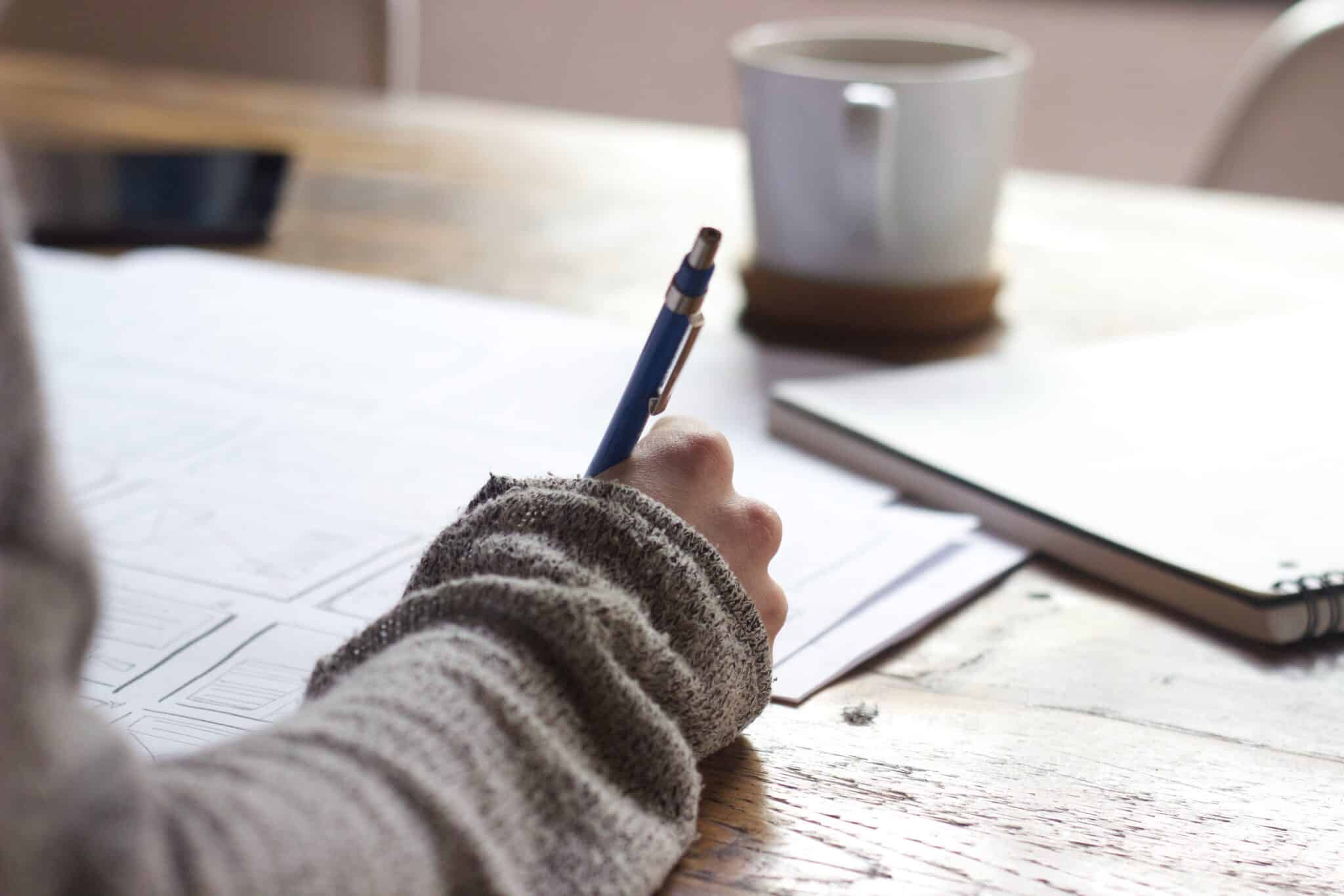 Person drawing something with a pen next to a cup of coffee