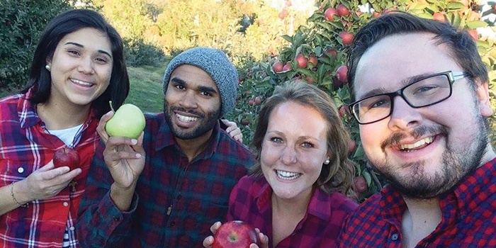 Four people smiling to the camera, 3 holding apples