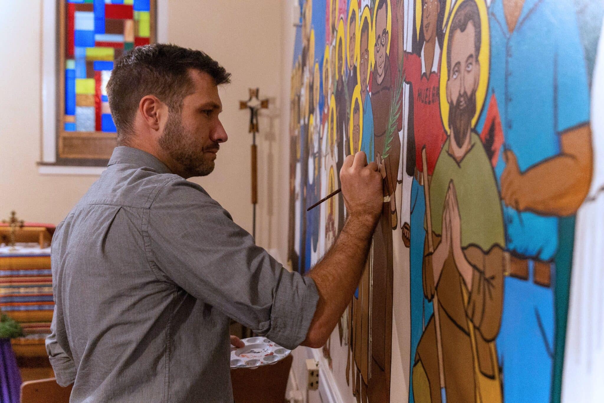 Jeremy Alexander, an iconographer and second-year theology teacher at Detroit Cristo Rey High School, is pictured Dec. 18, 2023, with a mural he created on a wall of the school's small chapel. Painted in his signature Byzantine style, the mural features 37 diverse saints, artists, poets, civil rights leaders and human rights activists. (OSV News photo/Steven Stechschulte, special to the Detroit Catholic)