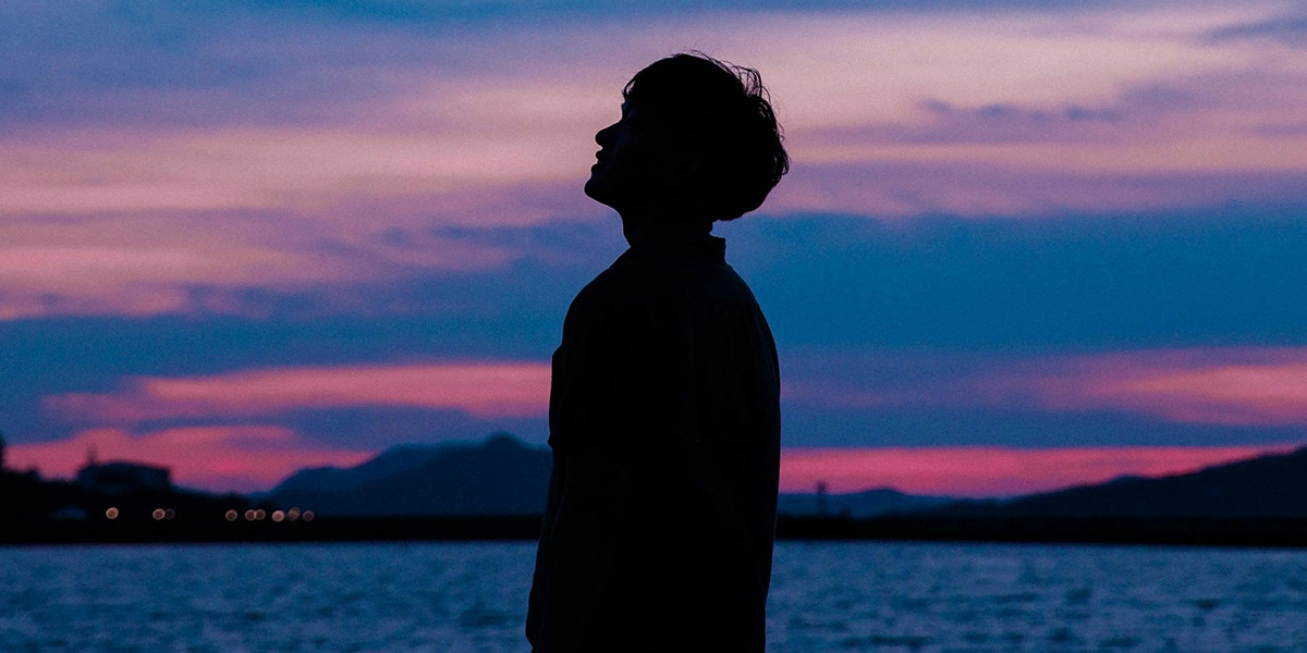 young man looking at the sky at night while standing by a lake.