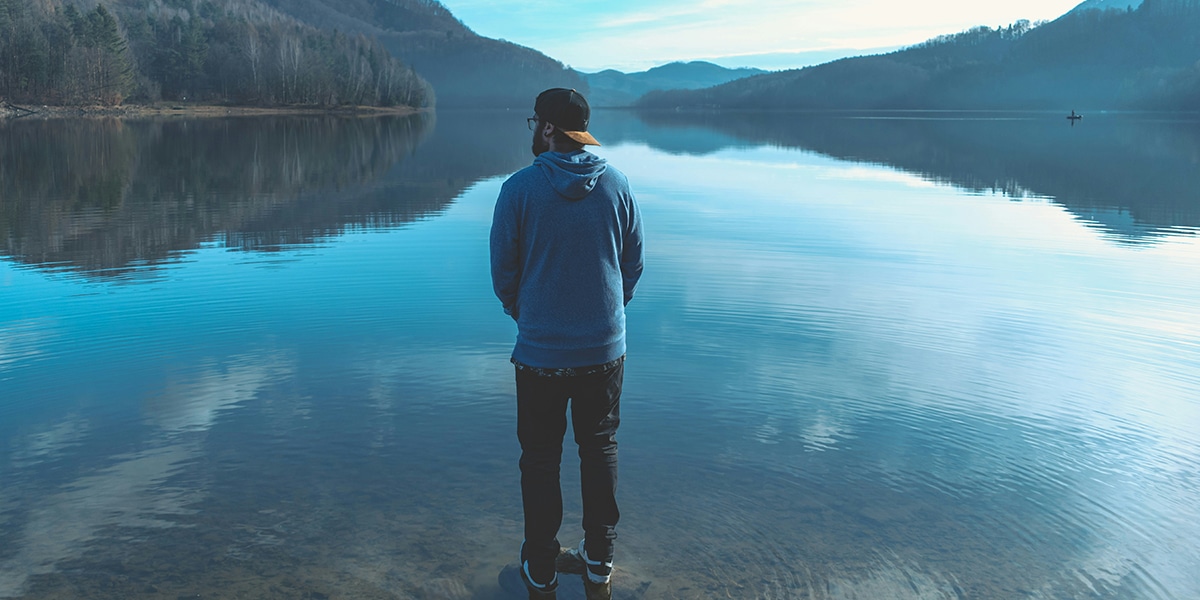 man standing by a lake and enjoying the view of the mountains.