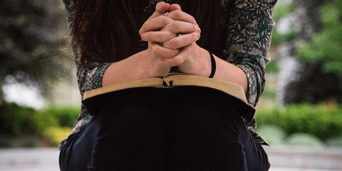 woman sitting down and praying with folded hands over a Bible.