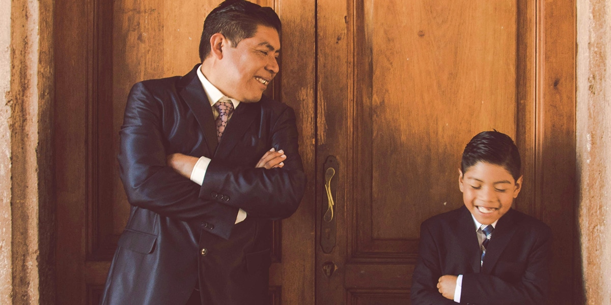 father and son enjoying a laugh while wearing suits and leaning against a door.