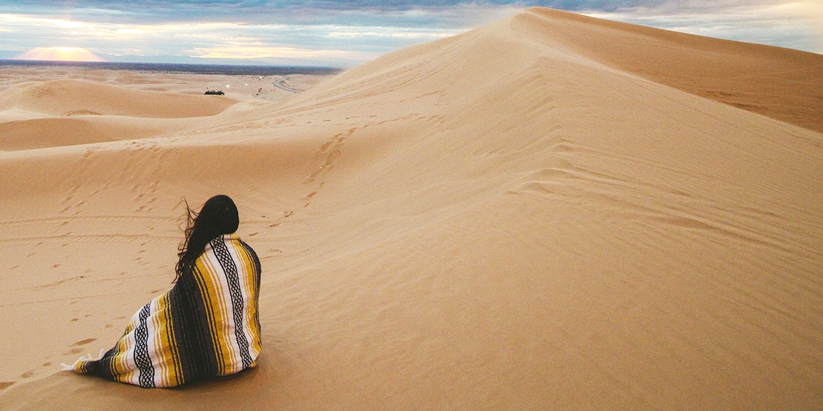 person sitting in the dessert, surrounded by sand dunes, covered by a blanket.