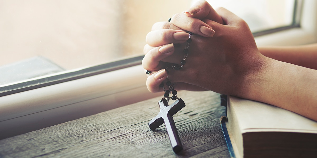 folded hands in prayer holding a rosary with a cross.