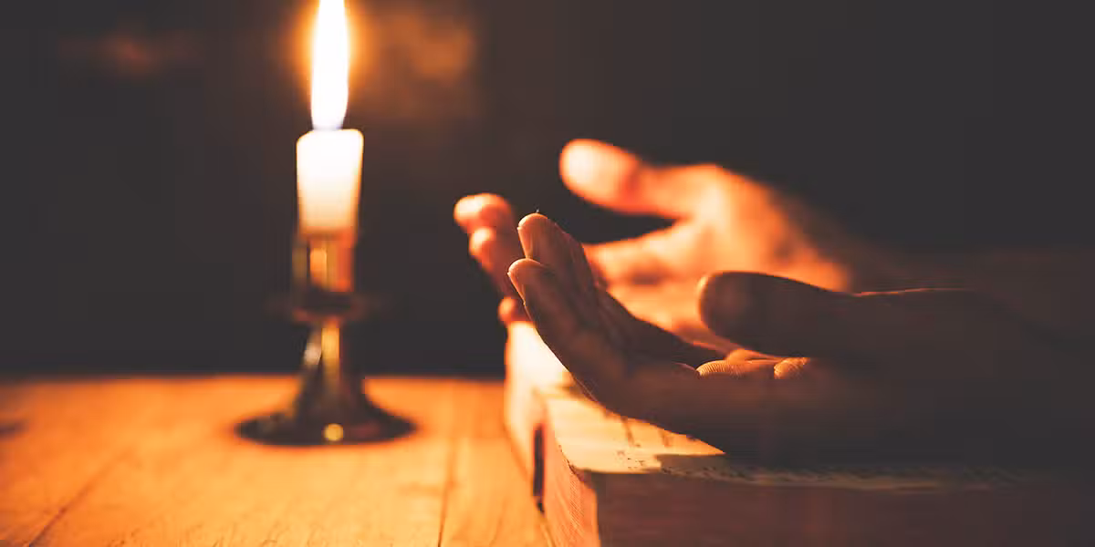 Hands open on a Bible in prayer next to a candle.