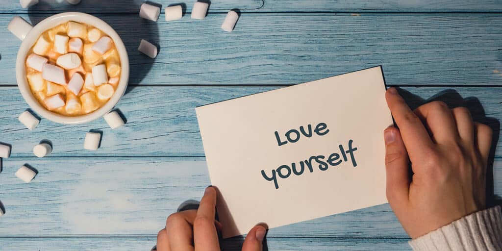 hands holding a note with the words "love yourself" written on it next to a cup of coffee
