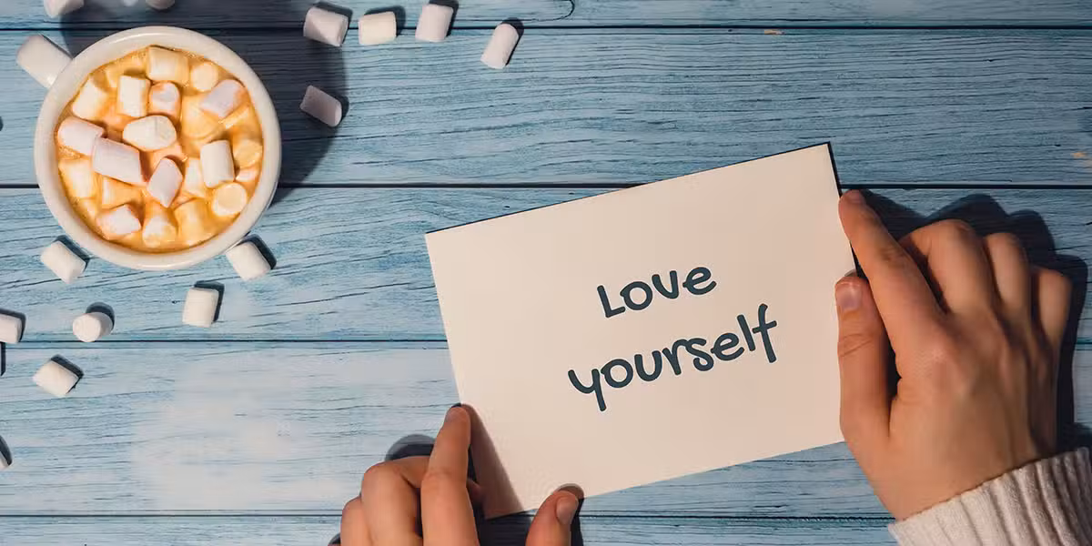 hands holding a note with the words "love yourself" written on it next to a cup of coffee