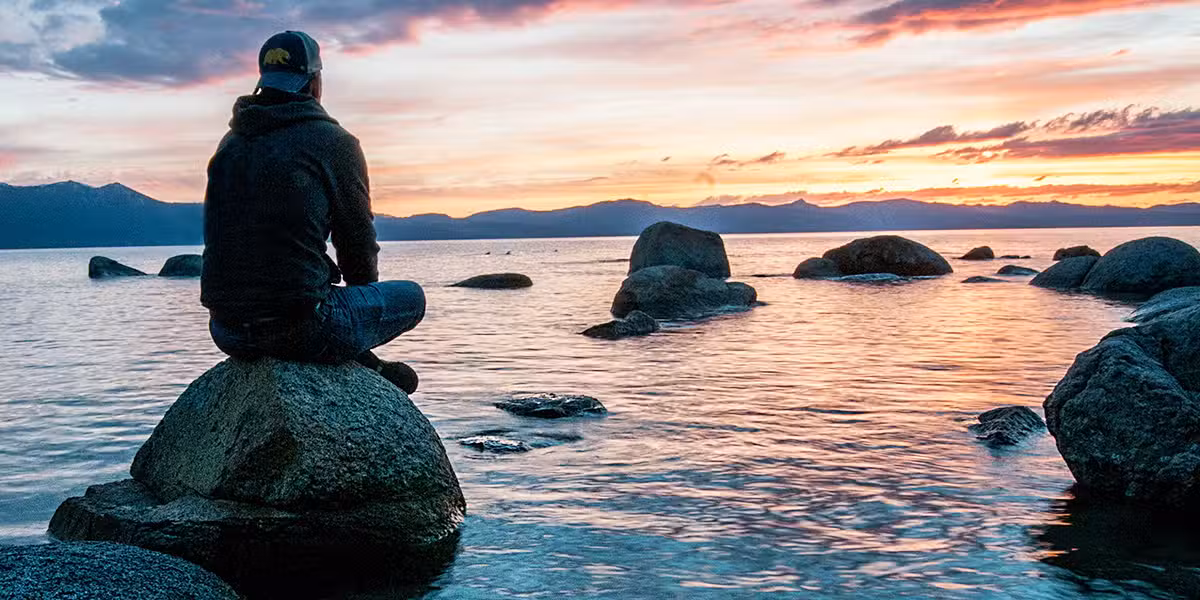 embrace silence and solitude; man sitting on a rock in the water, watching a sunset; Keegan Houser/Unsplash