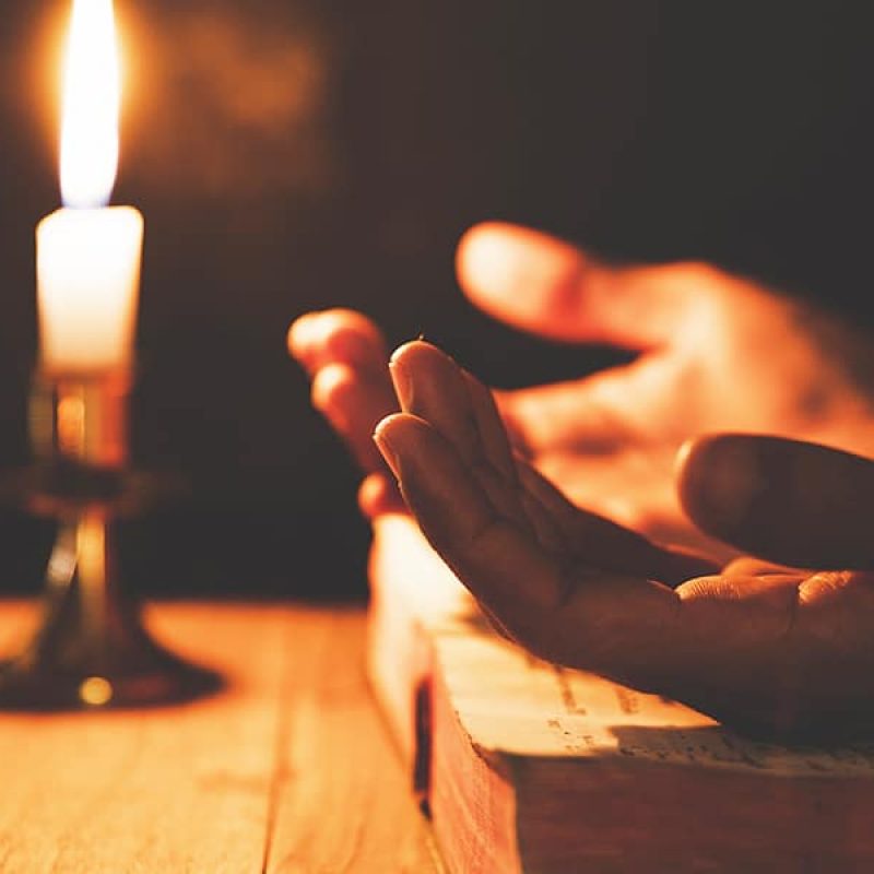 Hands open on a Bible in prayer next to a candle.