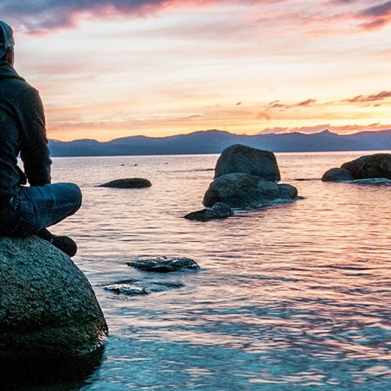 embrace silence and solitude; man sitting on a rock in the water, watching a sunset; Keegan Houser/Unsplash