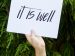 hand is holding a sign with the words "It Is well"