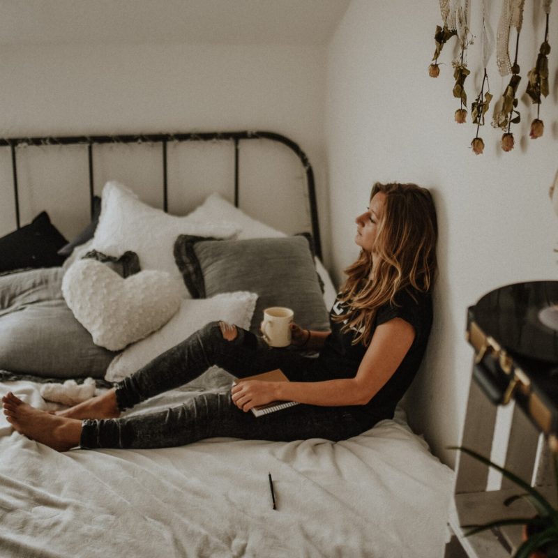Person sitting with back against the wall on the bed holding coffee