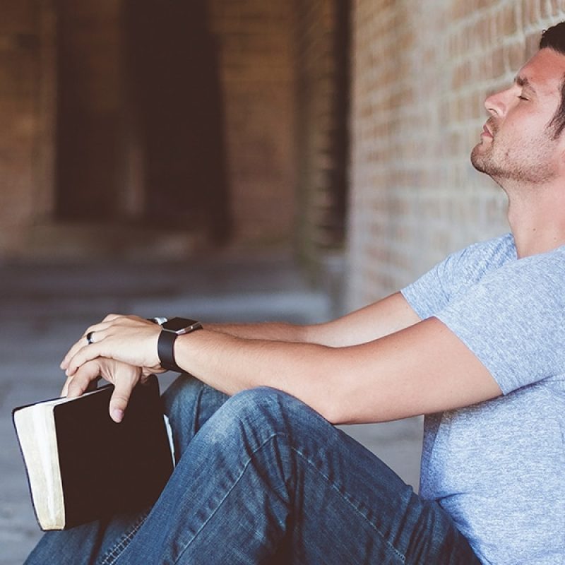 Man praying with his eyes closed while holding a Bible and leaning against a wall