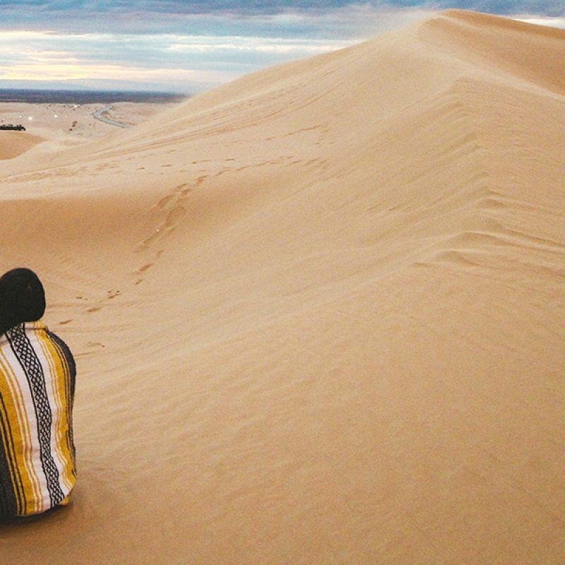 person sitting in the dessert, surrounded by sand dunes, covered by a blanket.