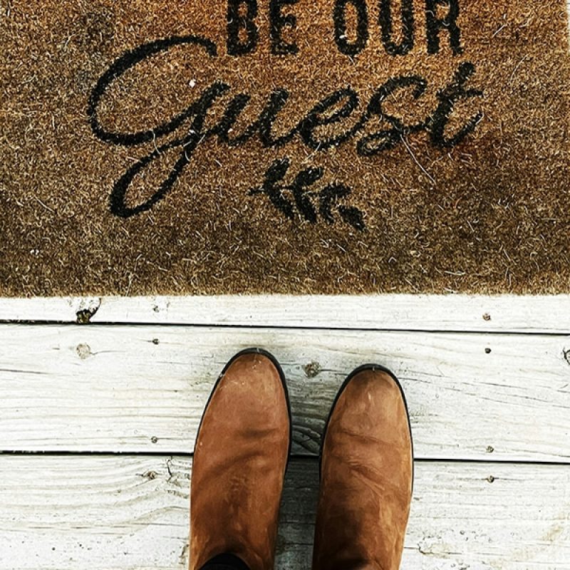 shoes standing in front of a welcome mat with the words "Be Our Guest"